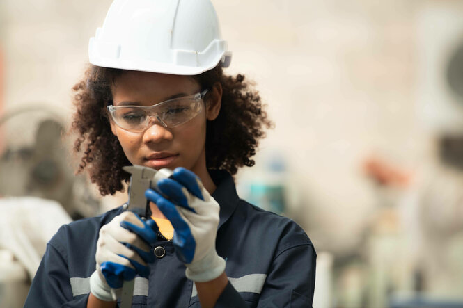 Eine junge Frau mit Bauhelm inspiziert und repariert Teile eines Schweißroboters. Symbolbild