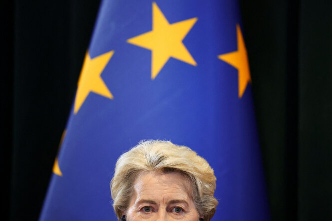 Ursula von der Leyen guckt kurz über den unteren Bildanschnitt, im Hintergrund die Fahne der EU