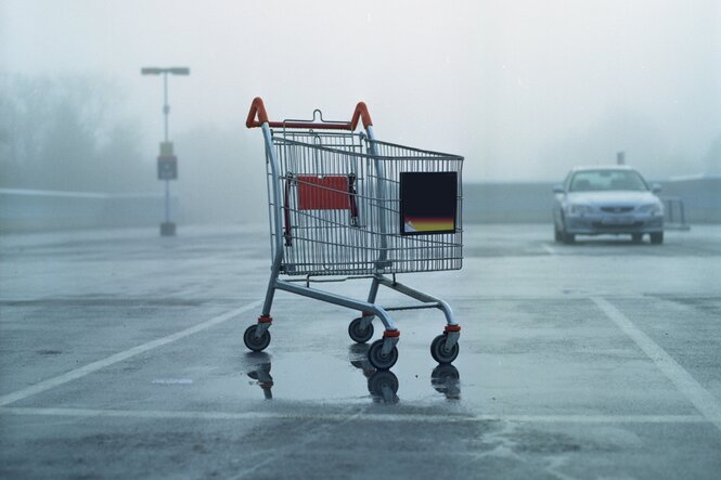 Leerer Einkaufswagen steht auf einem tristen Parkplatz, im Hintergrund ein Auto, neblige Stimmung