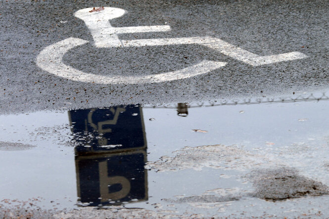 Ein Piktogram mit einem Strichmännchen im Rollstuhl auf dem Boden weist auf einen Behindertenparkplatz hin, in einer Pfütze darunter spiegelt sich außerdem das entsprechende Hinweisschild