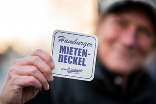 Mann hält einen Bierdeckel hoch auf dem steht: "Hamburger Mietedeckel"