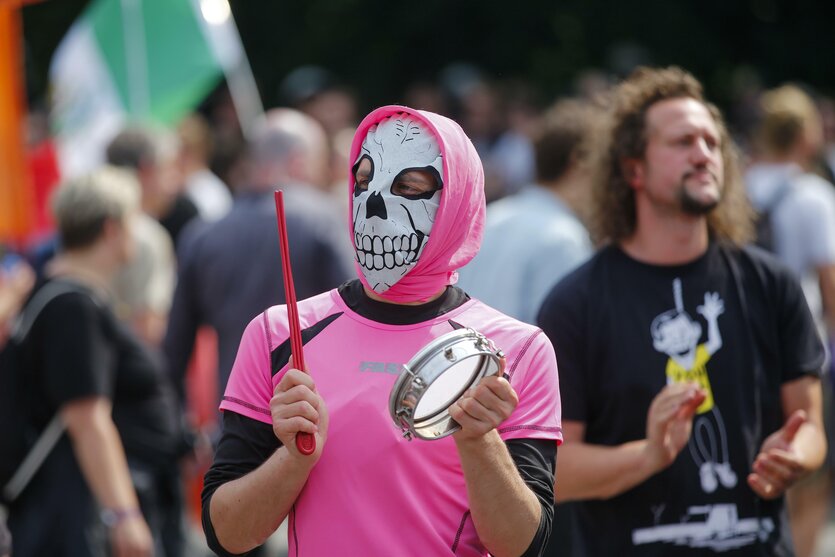 Eine Person, in einem pinken Anzug und mit Maske, schlägt auf ein Tamburin