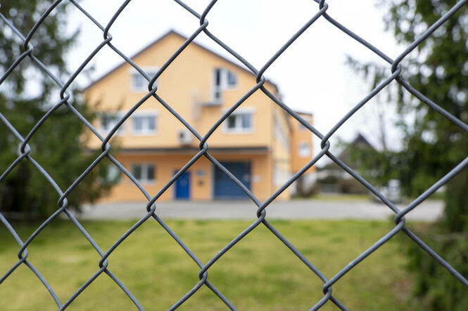 Hinter einem Maschendrahtzaun ist ein Haus unscharf zu erkennen