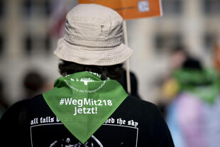 Eine demonstrierende Person mit einem Tuch, auf dem steht: " #Wegmit218 Jetzt!"