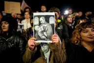 Eine Frau hält auf einer Protestaktion in Bukarest ein kleines Plakat in die Kamera. Es zeigt einem klassischen Gemälde nachempfunden Wladimir Putin, der und Calin Georgescu als Baby in den Armen wiegt