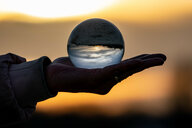 Ein Frau hält am Strand in Surendorf eine Glaskugel in der Hand, in der sich die Umgebung und der Sonnenuntergang an der Eckernförder Bucht spiegeln.