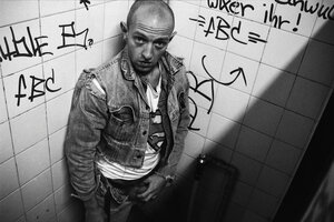 Schwarzweiß Foto eines Mannes in einer Toilette mit beschmierter Wand. Der Mann trägt eine Jeansjacke, hat seine Hose aufgeknöpft und fasst sich an den Penis