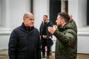 Olaf Scholz und Wolodomyr Selenskyj stehen in dicken Jacken vor einem Gebäude und sprechen miteinander. Selenskyi hebt fragend die Arme hoch