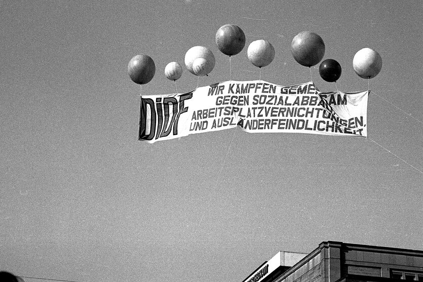 Luftballons tragen ein Banner vor freiem Himmel in die Höhe, in Schwarzweiß.