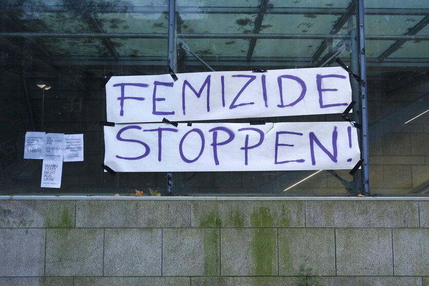 Ein weißes Banner mit lilaner Schrift "Femizide stoppen", das über einer Mauer hängt