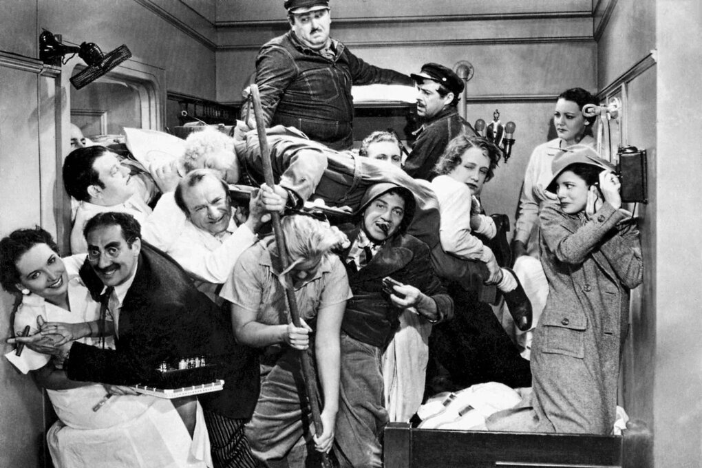 Szene aus einem Schwarz-Weiß-Film: Unzählige Gäste stapeln sich in einer Schiffskabine übereinander. Ein Mann hält in der Bildmitte einen Wischmob in der Hand. Einer der Marx Brothers hält links eine Zigarre zwischen den Fingern und lacht.