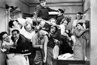 Szene aus einem Schwarz-Weiß-Film: Unzählige Gäste stapeln sich in einer Schiffskabine übereinander. Ein Mann hält in der Bildmitte einen Wischmob in der Hand. Einer der Marx Brothers hält links eine Zigarre zwischen den Fingern und lacht.