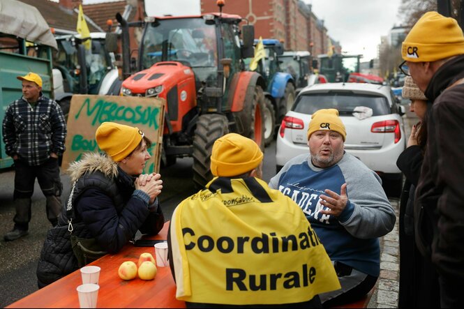 Menschen sitzen an einem Holztisch auf der STraße, Bauern mit Traktoren protestieren dahinten