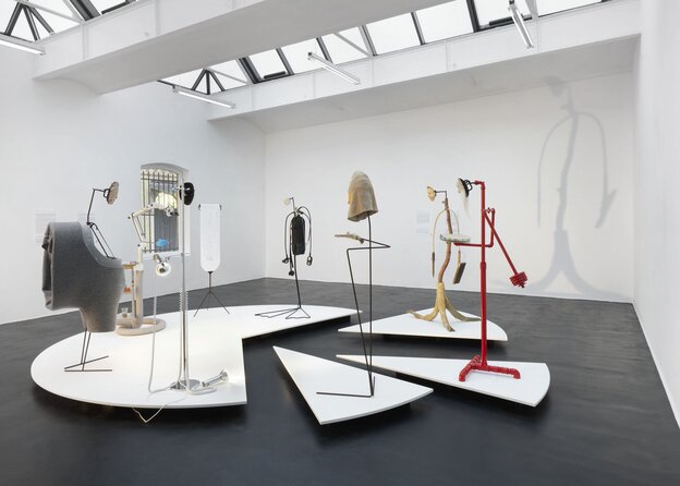Blick in einen Ausstellungsraum mit weißen Wänden und dunkelgrauem Boden. Eine Gruppe von Skulpturen steht auf einem Tortendiagram-förmigen Plateau. Die Skulpturen setzten sich aus Stahl, Stoff- und Plastikelementen zusammen und deuten abstrahierte Körper