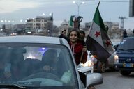 Ein Mädchen hängt mit ihrem Oberkörper aus einem Auto. In ihrer linken Hand hält sie die Fahne der syrischen Oppositionsbewegung.