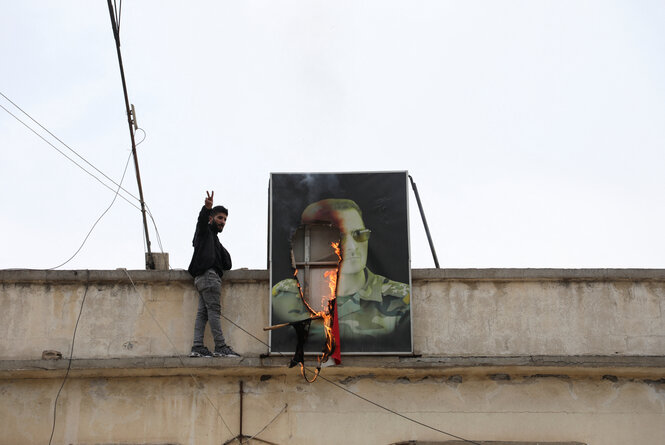 Auf einem Dach brennt ein Bild von Baschar al-Assad, daneben zeigt ein Mann das Victory-Zeichen