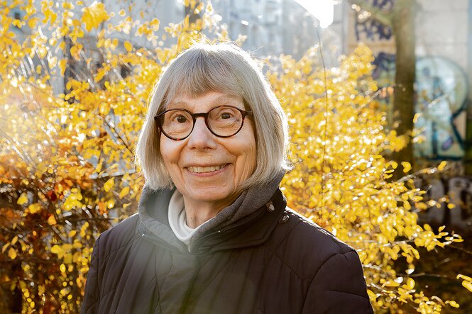 Eine ältere Frau, Gretchen Dutschke Klotz, steht draußen im Sonnenlicht und lächelt in die Kamera, im Hintergrund schimmert Herbstlaut in goldgelben Farben.