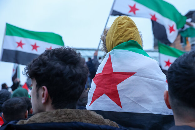 Der Rücken eines jungen Mannes und eines Kindes, das in eine syrische Flagge gehüllt ist, sind in einer Menschenmenge mit weiteren syrischen Fahnen