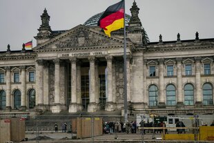 Das Reichstagsgebäude mit großer Deutschlandfahne, davor stehen Bauzäune und eine Besucherschlange