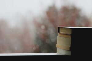 Ein Stapel Bücher vor einem mit Regentropfen benetzten Fenster, draußen sieht man Bäume mit gelb-braunen Blättern und grauen Himmel