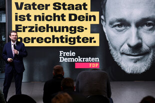 Ein Mann steht mit einem Mikrofon vor einem Wahlkampf-Plakat der FDP und spricht zu seinem Publikum