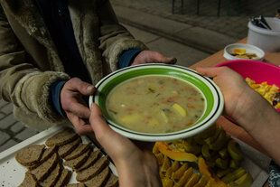 Zwei Hände halten einen Teller voll Suppe und geben ihn in zwei andere Hände