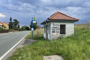 Der Himmel ist grau. Ein Bushaltestellenhäuschen mit Haltestellenschild steht mit rotem Ziegeldach am Schnellstraßenrand. An der grauen Fassade steht mit Grafitti gesprüht 