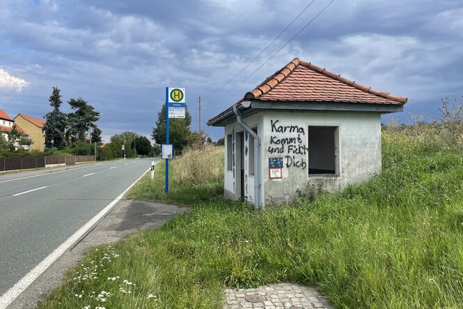 Der Himmel ist grau. Ein Bushaltestellenhäuschen mit Haltestellenschild steht mit rotem Ziegeldach am Schnellstraßenrand. An der grauen Fassade steht mit Grafitti gesprüht "Karma kommt und F*ckt Dich"