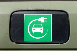 Schild einer Ladesäule. Es ist ein grünes Quadrat. Darin in weiß ein stilisiertes Auto von vorne. Um das Auto ist ein Kreis gelegt aus einem Kabel, an dessen Ende ein Steckdosenstecker ist.