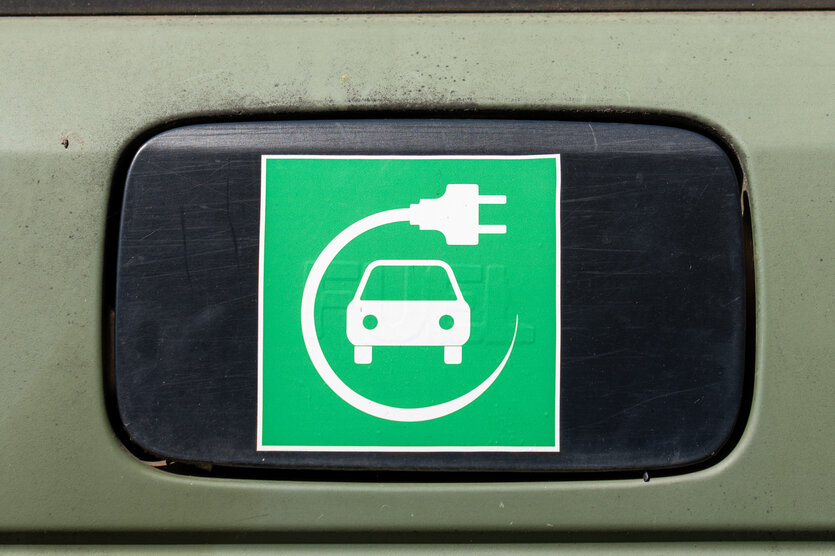 Schild einer Ladesäule. Es ist ein grünes Quadrat. Darin in weiß ein stilisiertes Auto von vorne. Um das Auto ist ein Kreis gelegt aus einem Kabel, an dessen Ende ein Steckdosenstecker ist.