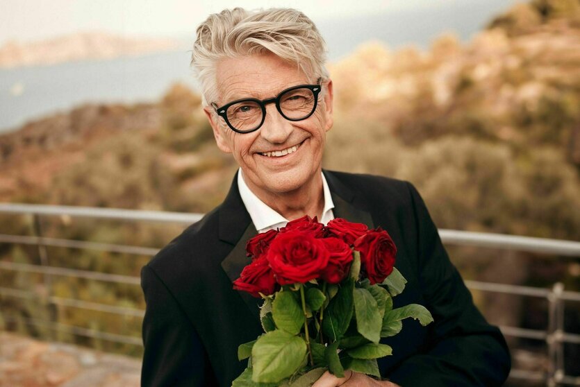 Ein älterer Mann mit einem Rosenstrauß lächelt in die Kamera. Er hat graues, volles Haar und trägt eine Brille mit dunklem, dicken Rahmen. Hinter ihm viel Landschaft in Orange-Tönen.