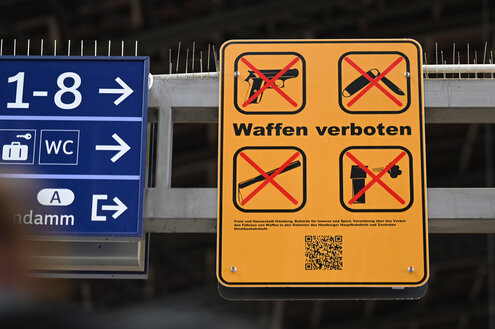 Ein Schild mit durchgeschtrichenen Symbolen weist an einem Bahnhof auf das Waffenverbot hin