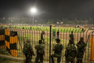 Soldaten stehen vor einem Zaun, dahinter findet ein Fußballspiel statt