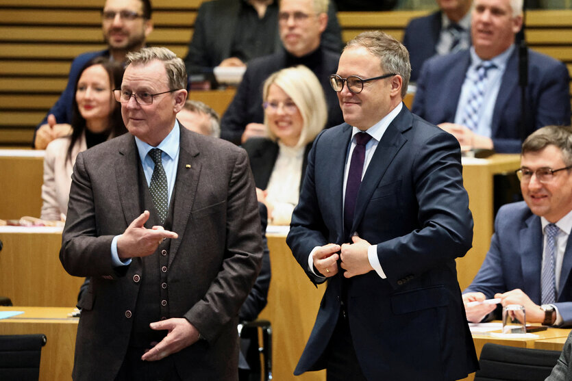 Bodo Ramelow und Mario Voigt stehen im Sitzungssaal des thüringischen Landtags nebeneinander, Ramelow zeigt auf Voigt, Voigt knöpft sich das Jacket