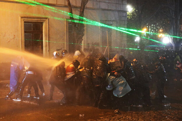 Eine Gruppe von Menschen steht eng in einer Traube zusammen, um sich vor dem Strahl eines Wasserwerfers zu schützen, über ihenen sind die grünen Strahlen eines Laserpointers zu sehen
