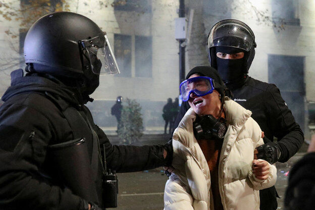 Eine Frau mit Schutzbrille wird von zwei Polizisten in schwarzer Montur festgehalten, sie sperrt den Mund auf
