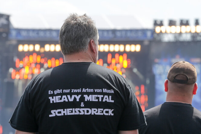 Ein Besucher des Wacken Open Air mit einem T-Shirt Aufdruck „Es gibt nur zwei Arten von Musik / Heavy Metal & Scheissdreck“ schaut auf Bühne