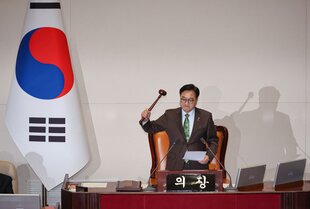 Der Sprecher der Nationalversammlung Woo Won-shik schwingt mit seiner rechten Hand einen Hammer, in der linken Hand hält er ein Blatt Papier. Er steht hinter seinem Platz, neben ihm ist eine südkoreanische Fahne platziert