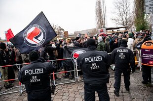 Im Vordergrund mehrere Bereischaftspolizisten, im Hintergrund eine Absperrung, dahinter viele Demonstrierende, einer schwingt eine große Antifa-Fahne