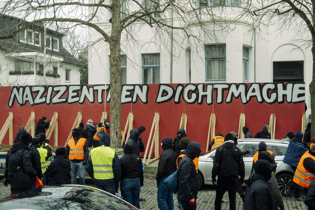 Vermummte vor einer roten Wand mit der Aufschrift 