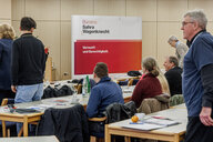 Menschen sitzen an Tischen und stehen davor, vorne steht eine rot-weiße Stellwand mit Schrift