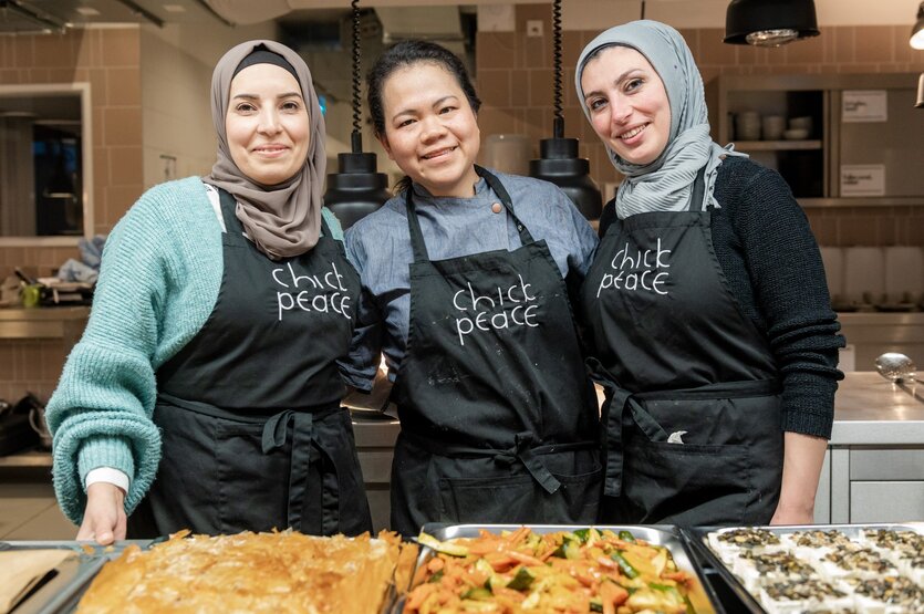 Drei Frauen stehen vor Blechen mit Essen in einer Küche.