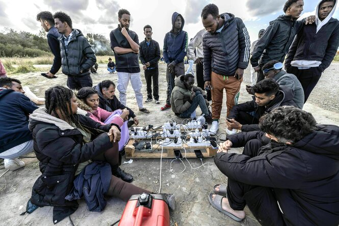 MigrantInnen sitzen in warmen Jacken im Kreis um eine Holzpalette, auf der viele Ladegeräte und Smartphones liegen. Der Strom dafür kommt von einem Generator