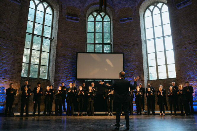 Ein Chor steht unter drei hohen Kirchenfenstern. Vor dem Chor steht ein Dirigent und hebt die Hände. Die Beteiligten sind schwarz gekleitet, hinter ihnen ist am Fenster in der Mitte eine unbespielte Videoleinwand zu sehen.