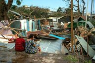 Zwei Frauen sitzen vor ihrem völlig zerstörten Haus, Wellblech, Holzltten und umgestürzte Bäume liegen auf dem Grundstück.