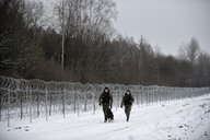 Zwei Grenzbeamte und ein Wachhund laufen entlang eines Grenzzauns in verschneiter Landschaft