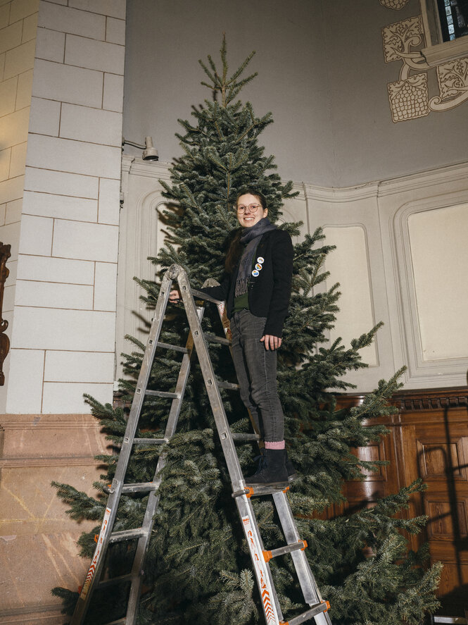 Eine junge Frau steht auf einer Leiter vor einem Tannenbaum, sie trägt einige Sticker am schwarzen Jackett