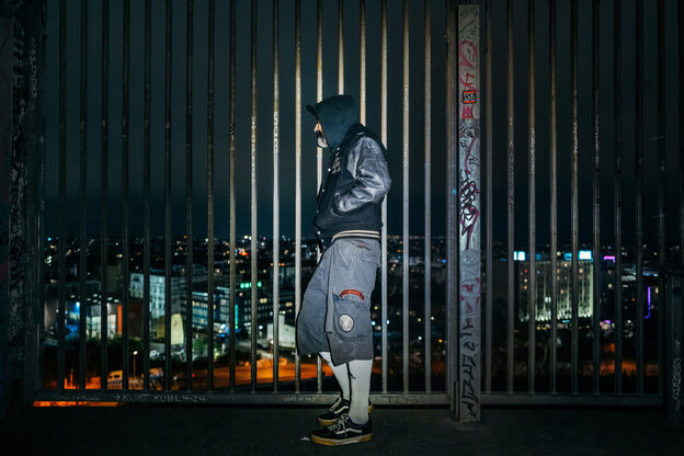 Ein Mann steckt die Hände in die Jacke und lehnt an eine Gitter, dahinter Lichter der Stadt