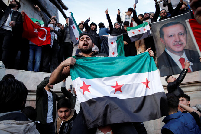 Syrische Geflüchtete jubeln in der Türkei; im Vordergrund weht eine Persodie neue syrische Flagge, umgebend im Hintergrund erhöht ist eine jubelnde Menge mit türkischer Flagge und Erdogan Porträt.