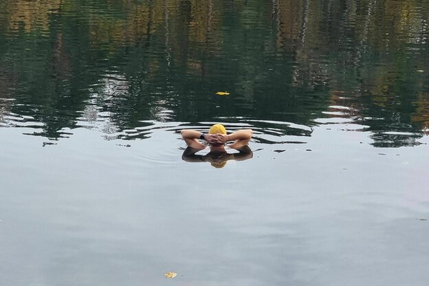 Eine Person steht im See, in dem sich Bäume spiegeln, und man sieht nur Kopf und Hände